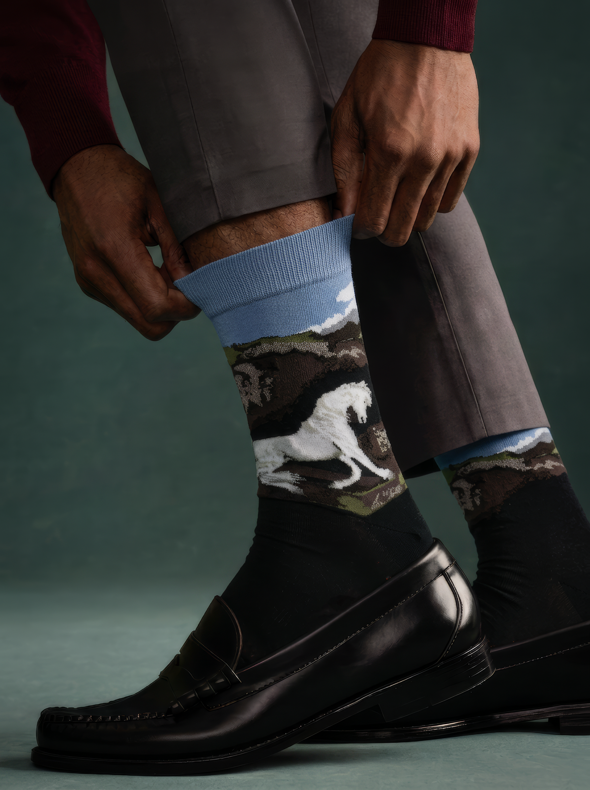 Person adjusting dark trousers to show charcoal ArtyGifts socks with pale blue cuffs and a black-and-white horse print, paired with black loafers.
