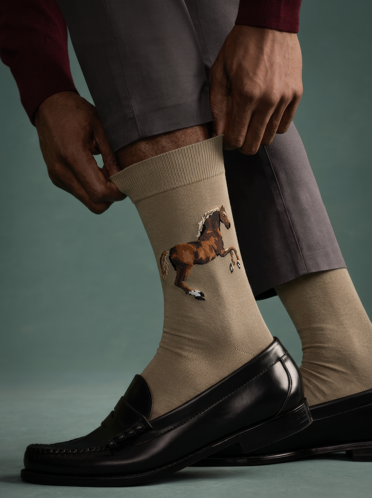 Close-up of a person adjusting taupe ArtyGifts socks featuring George Stubbs’s horse artwork, paired with grey trousers and black loafers.⠋