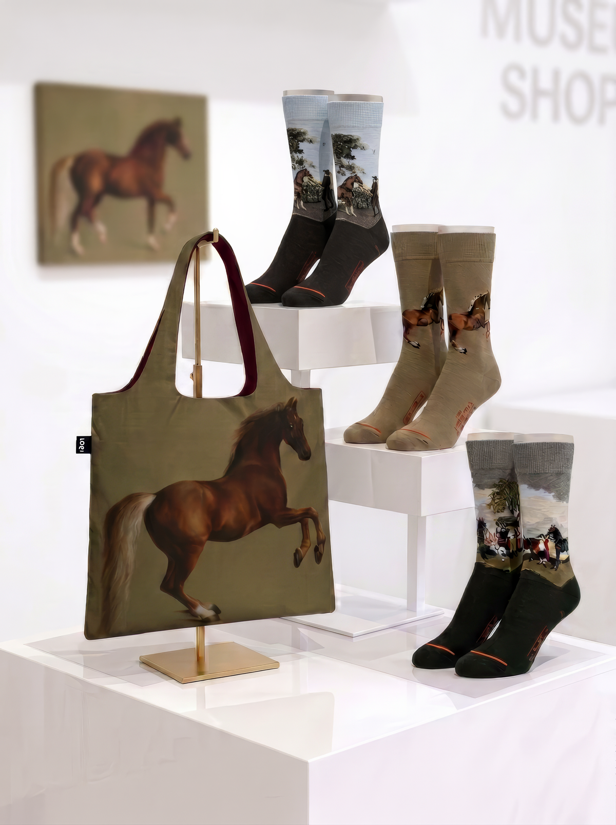 Three pairs of George Stubbs museum art socks and a matching foldable bag displayed on plinths in a museum shop.
