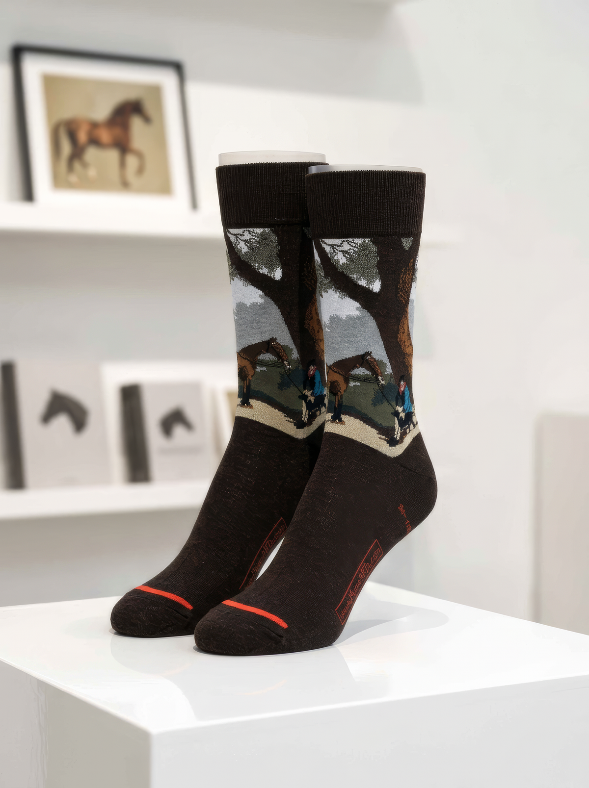 Pair of charcoal ArtyGifts socks with black cuffs and grey-white artwork from George Stubbs’s The Third Duke of Dorset’s Hunter, displayed on a white pedestal with art books behind them.

