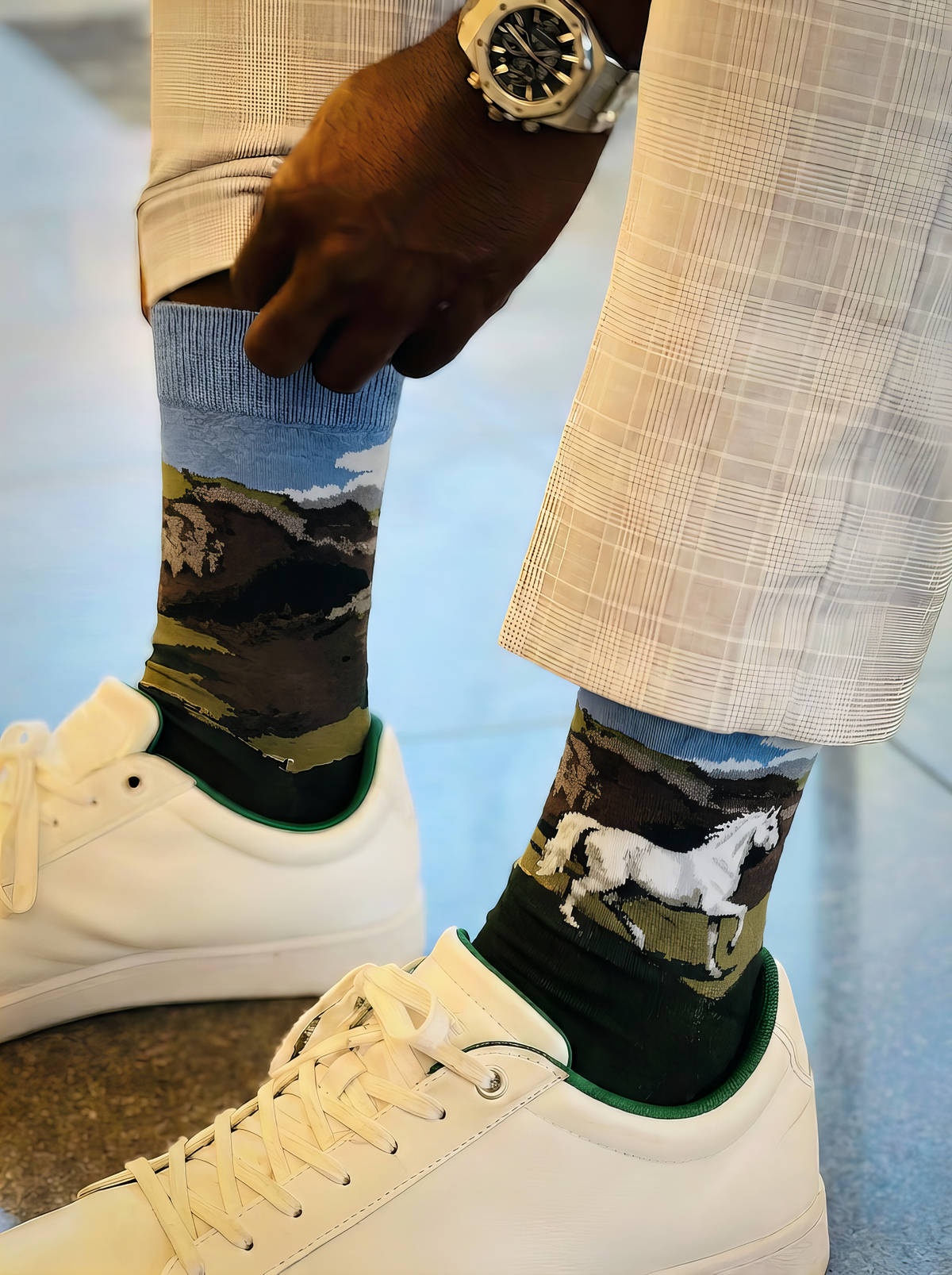 Close-up of a person adjusting charcoal ArtyGifts socks with pale blue cuffs and horse artwork, styled with white trousers.