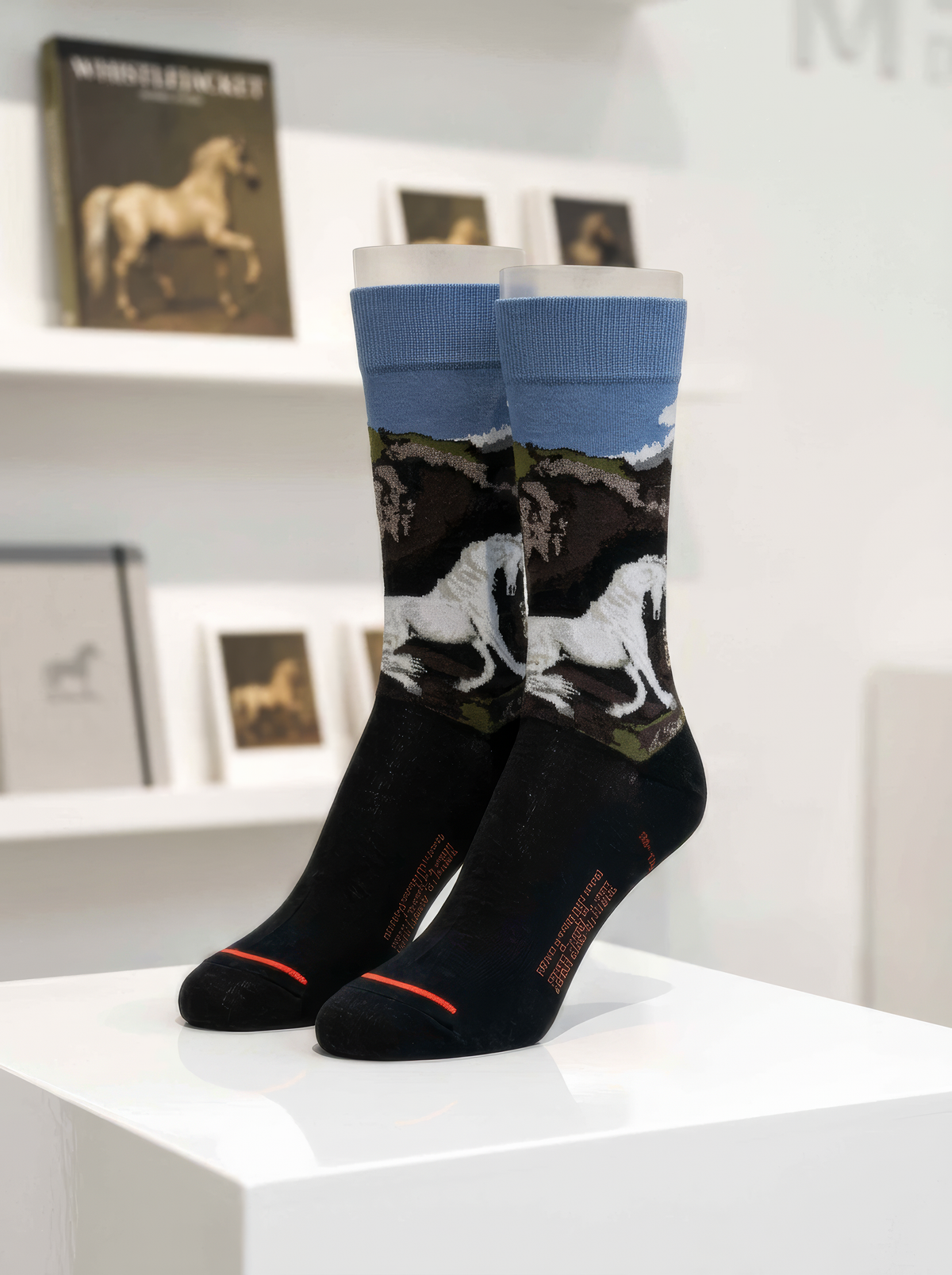 Pair of charcoal ArtyGifts socks with pale blue cuffs and black-and-white horse artwork, displayed on a white pedestal with horse prints behind them.