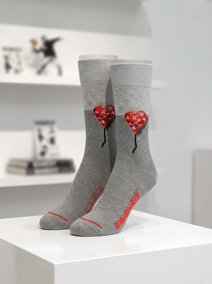 Pair of grey Bandaged Heart art socks with red heart motifs and black details, displayed upright on a white plinth in a gallery-style setting.

