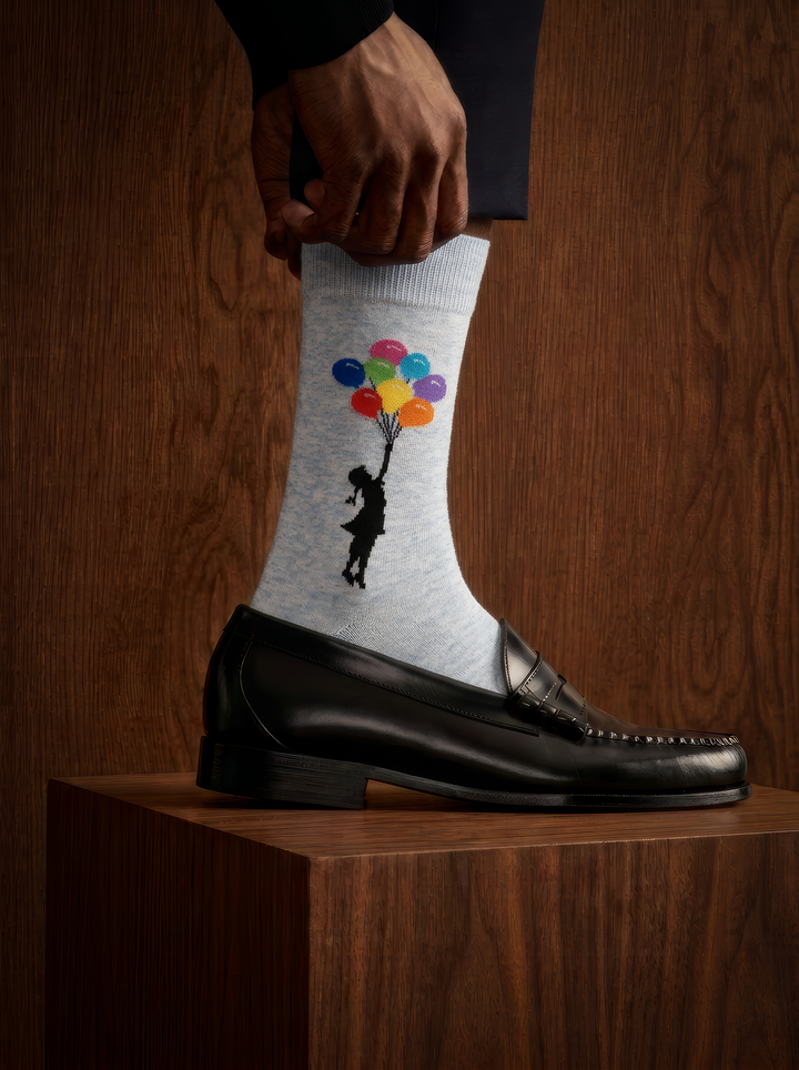 Close-up of a light grey Banksy-inspired balloon sock worn with a black leather shoe on a wooden plinth in a dimly lit setting.
