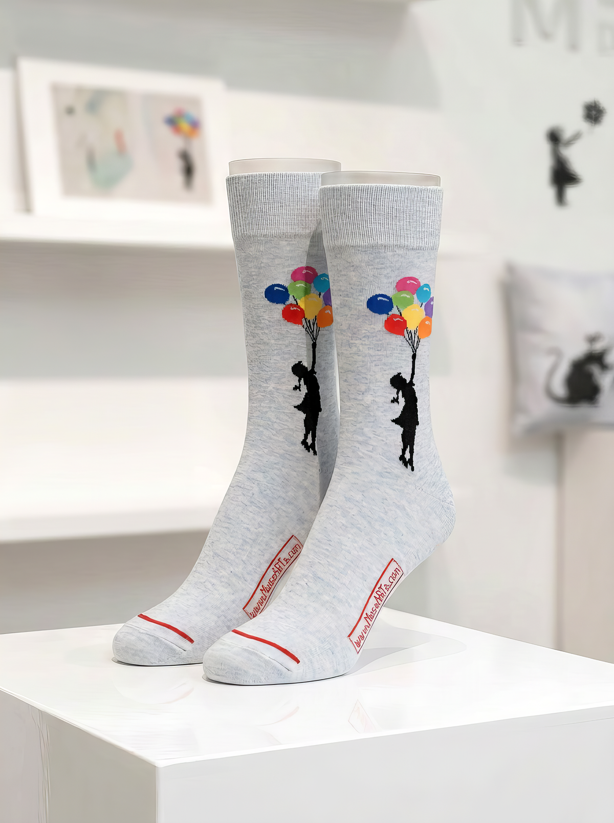 Pair of light grey Banksy-inspired art socks featuring black figures with colourful balloons, displayed on a white plinth in a gallery-style setting.
