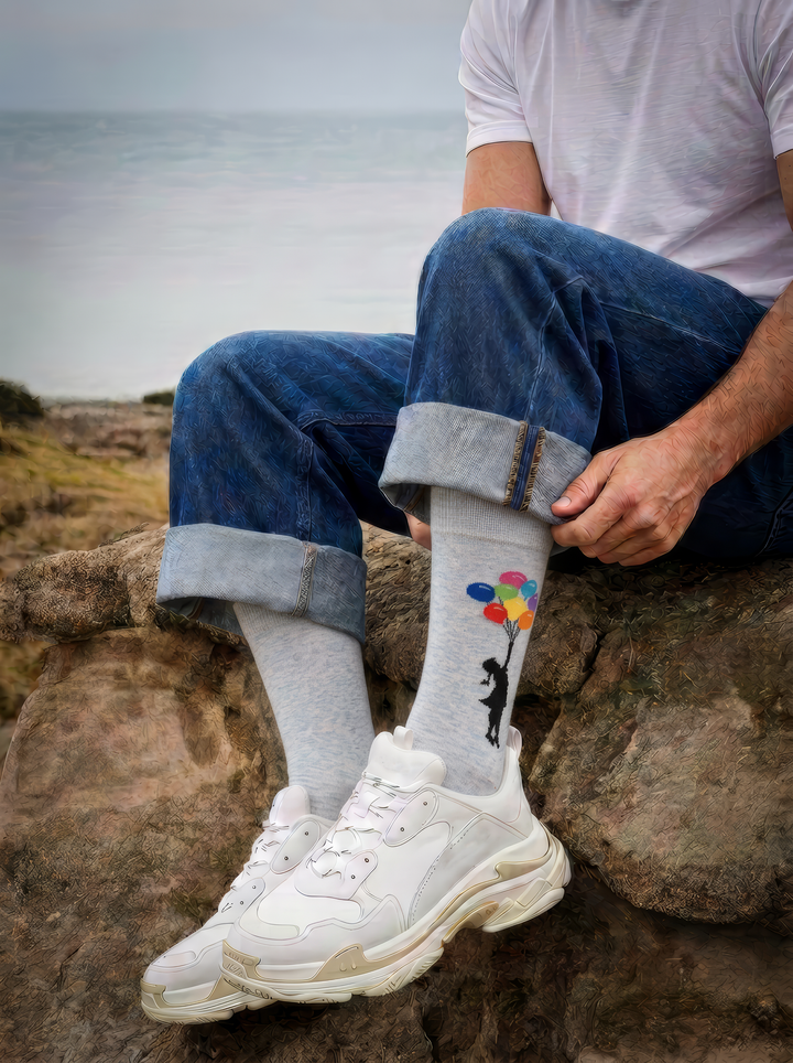 A seated person wears light grey Banksy-inspired balloon art socks with rolled blue jeans and white trainers outdoors.
