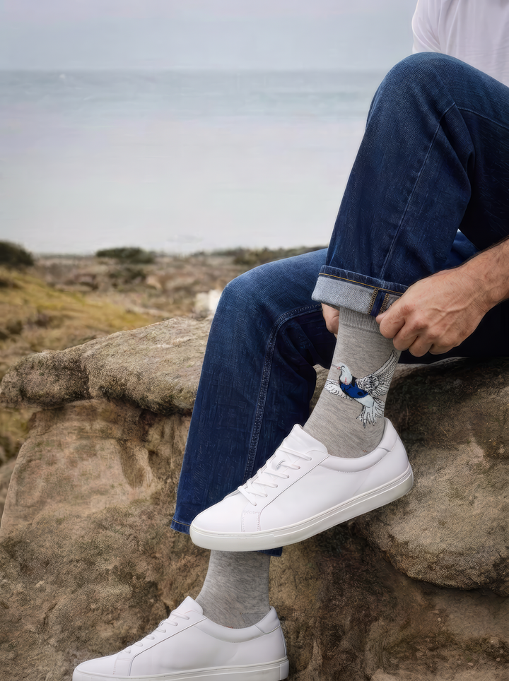 A seated person adjusts grey Banksy-inspired Peace Dove art socks, styled with rolled blue jeans and white trainers outdoors.
