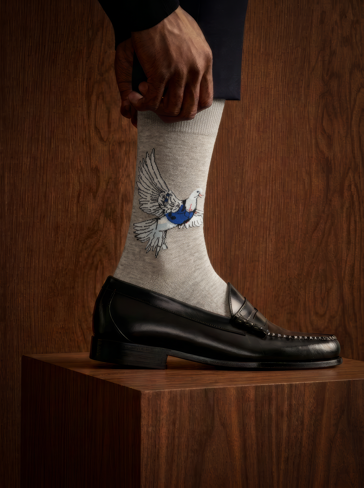 Close-up of a grey Banksy-inspired Peace Dove art sock worn with a black leather shoe resting on a wooden plinth in a dimly lit setting.
