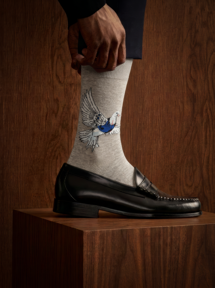 Close-up of a grey Banksy-inspired Peace Dove art sock worn with a black leather shoe resting on a wooden plinth in a dimly lit setting.
