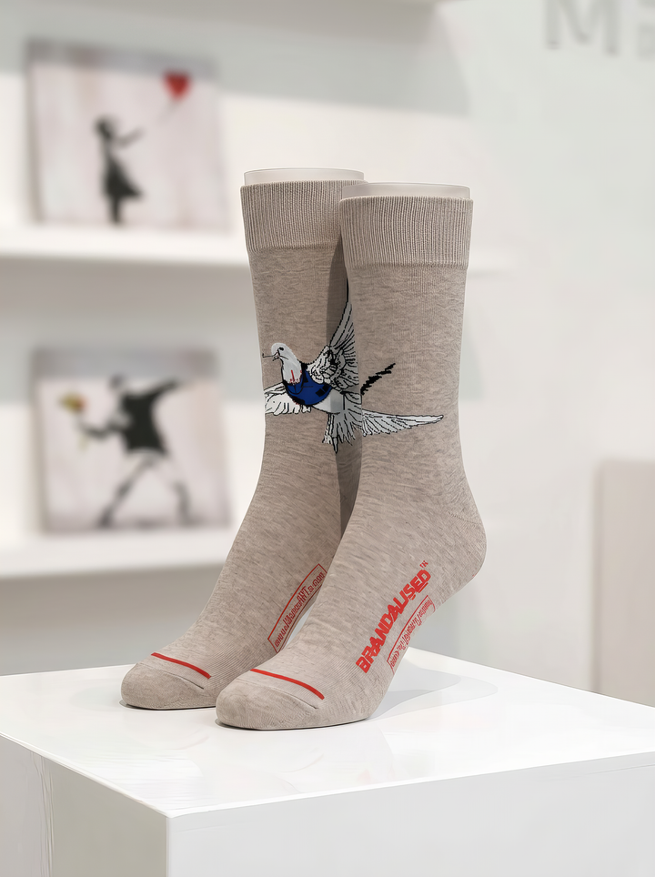Pair of warm grey Banksy-inspired Peace Dove art socks with a black and white dove motif and red accents, displayed on a white gallery-style plinth.
