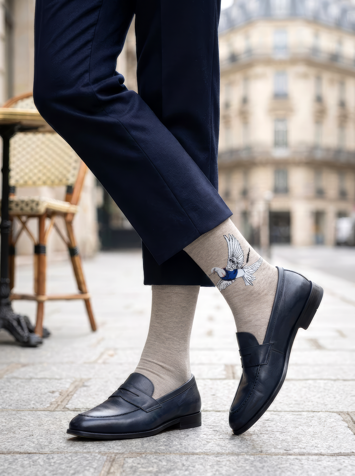 A seated person wears grey Banksy-inspired Peace Dove art socks with cropped dark trousers and black leather shoes in a bright interior.
