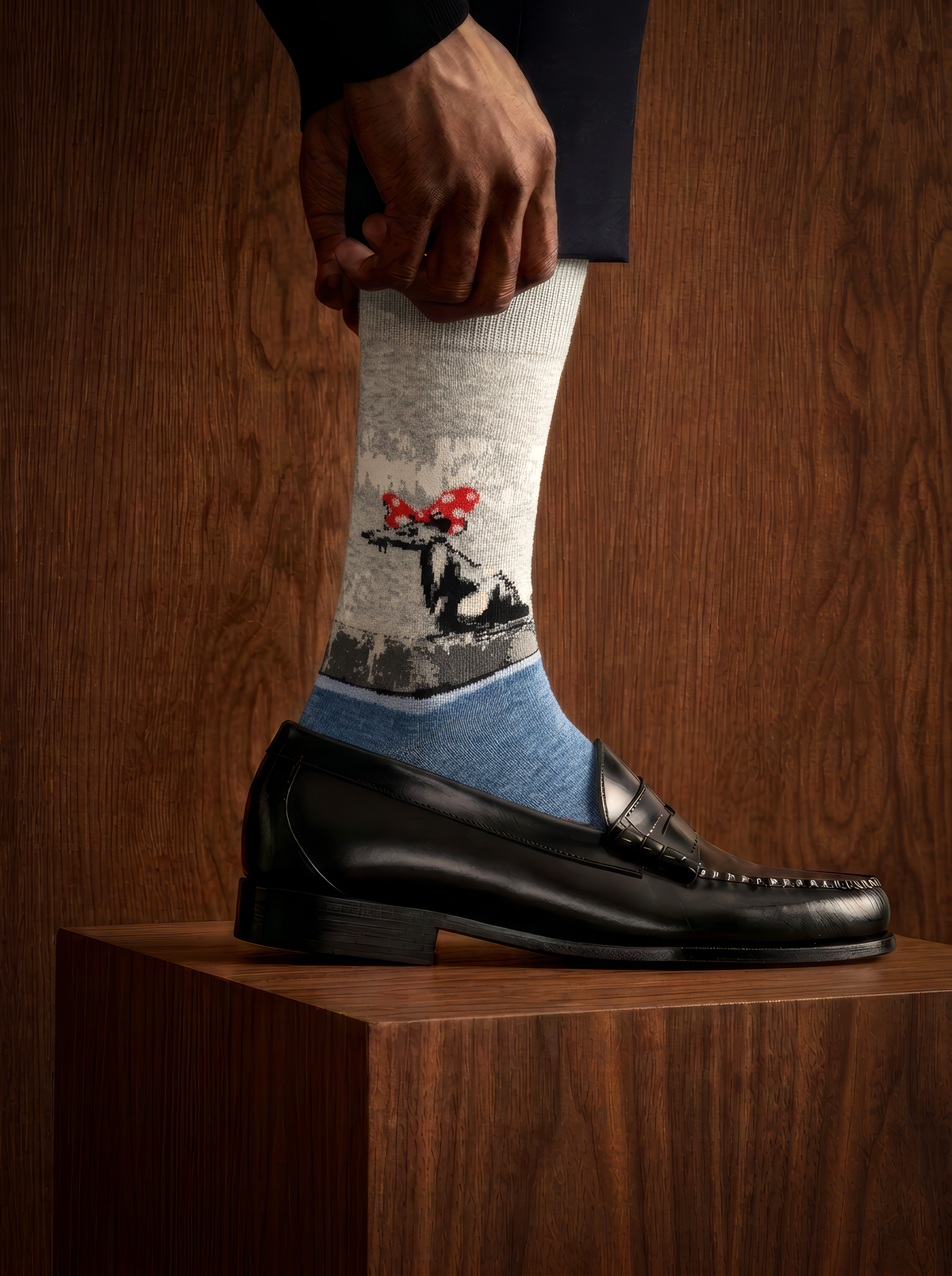 Close-up of a Banksy-inspired Polka Dot Rat art sock worn with a black leather shoe on a wooden plinth in a dimly lit setting.
