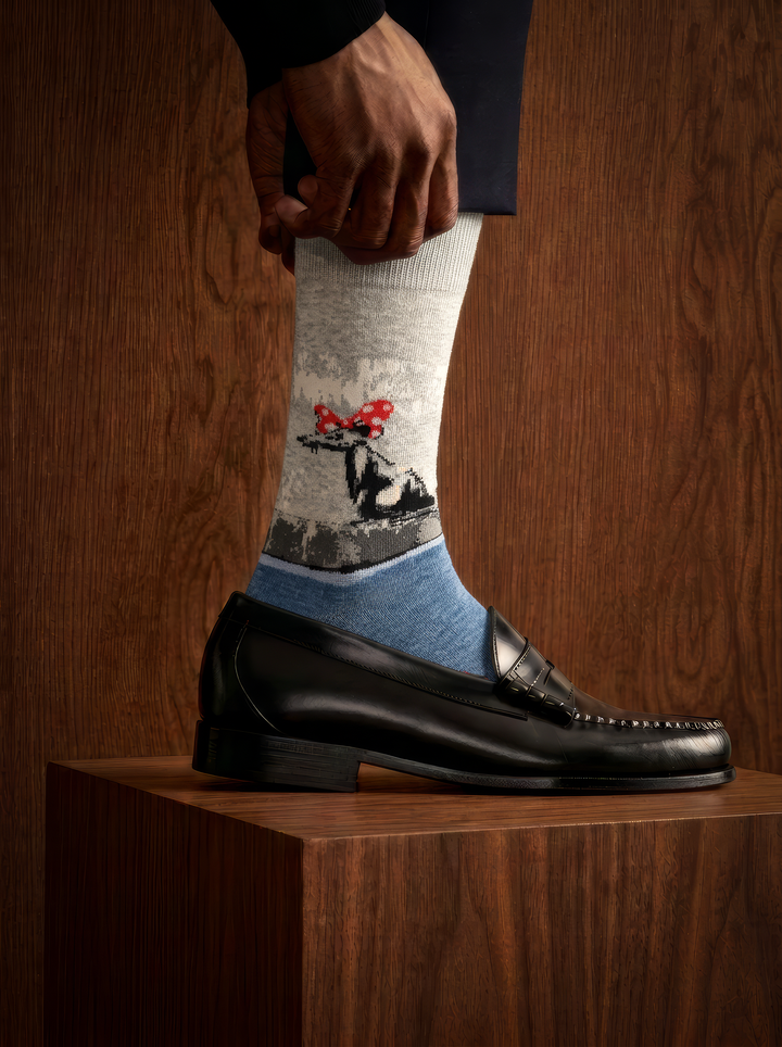 Close-up of a Banksy-inspired Polka Dot Rat art sock worn with a black leather shoe on a wooden plinth in a dimly lit setting.
