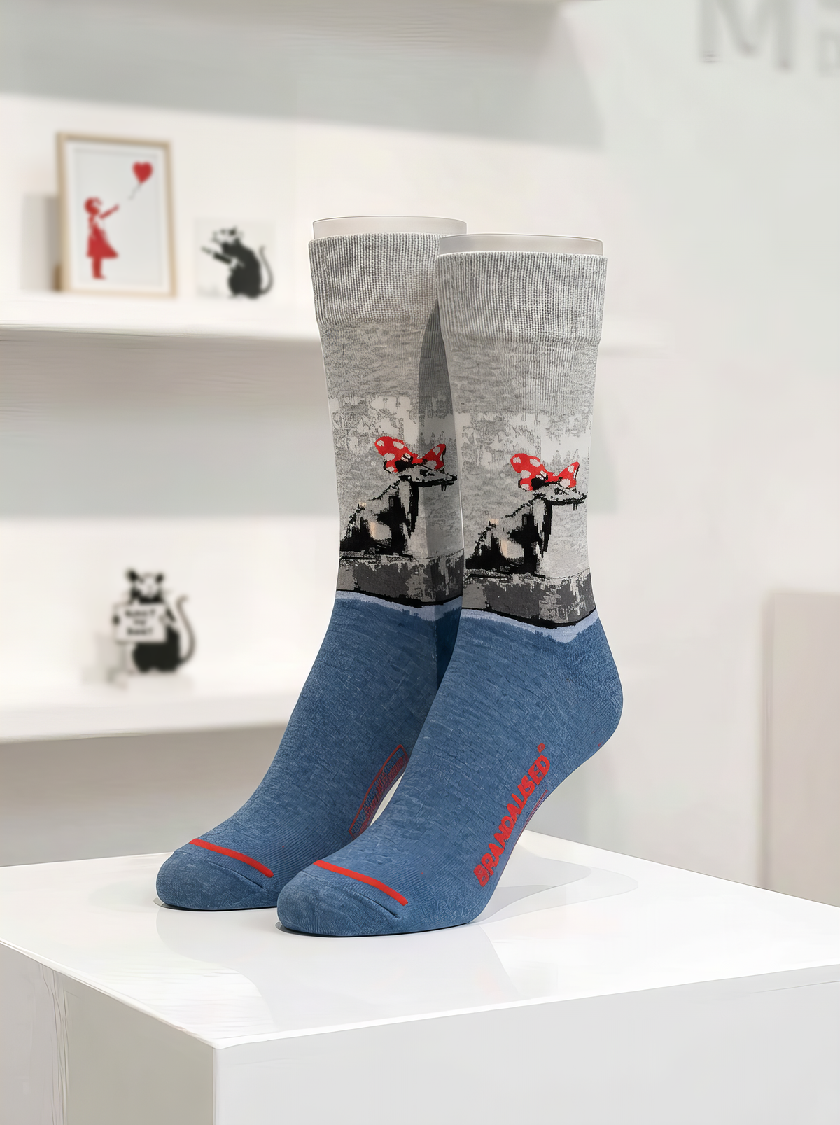 Pair of Banksy-inspired Polka Dot Rat art socks with grey illustrated legs, blue feet and red accents, displayed on a white gallery-style plinth.

