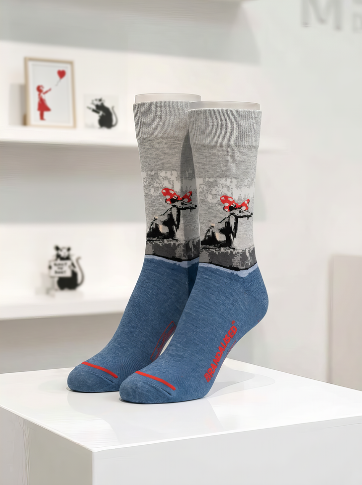 Pair of Banksy-inspired Polka Dot Rat art socks with grey illustrated legs, blue feet and red accents, displayed on a white gallery-style plinth.
