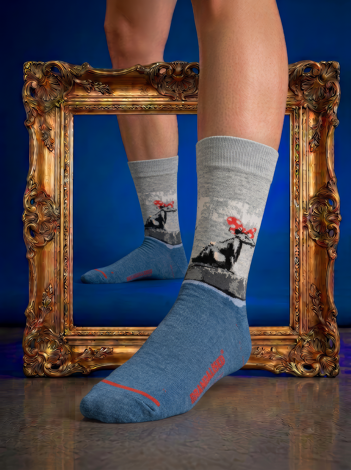 Banksy-inspired Polka Dot Rat art socks with grey legs and blue feet, worn beside an ornate gold frame against a deep blue background.
