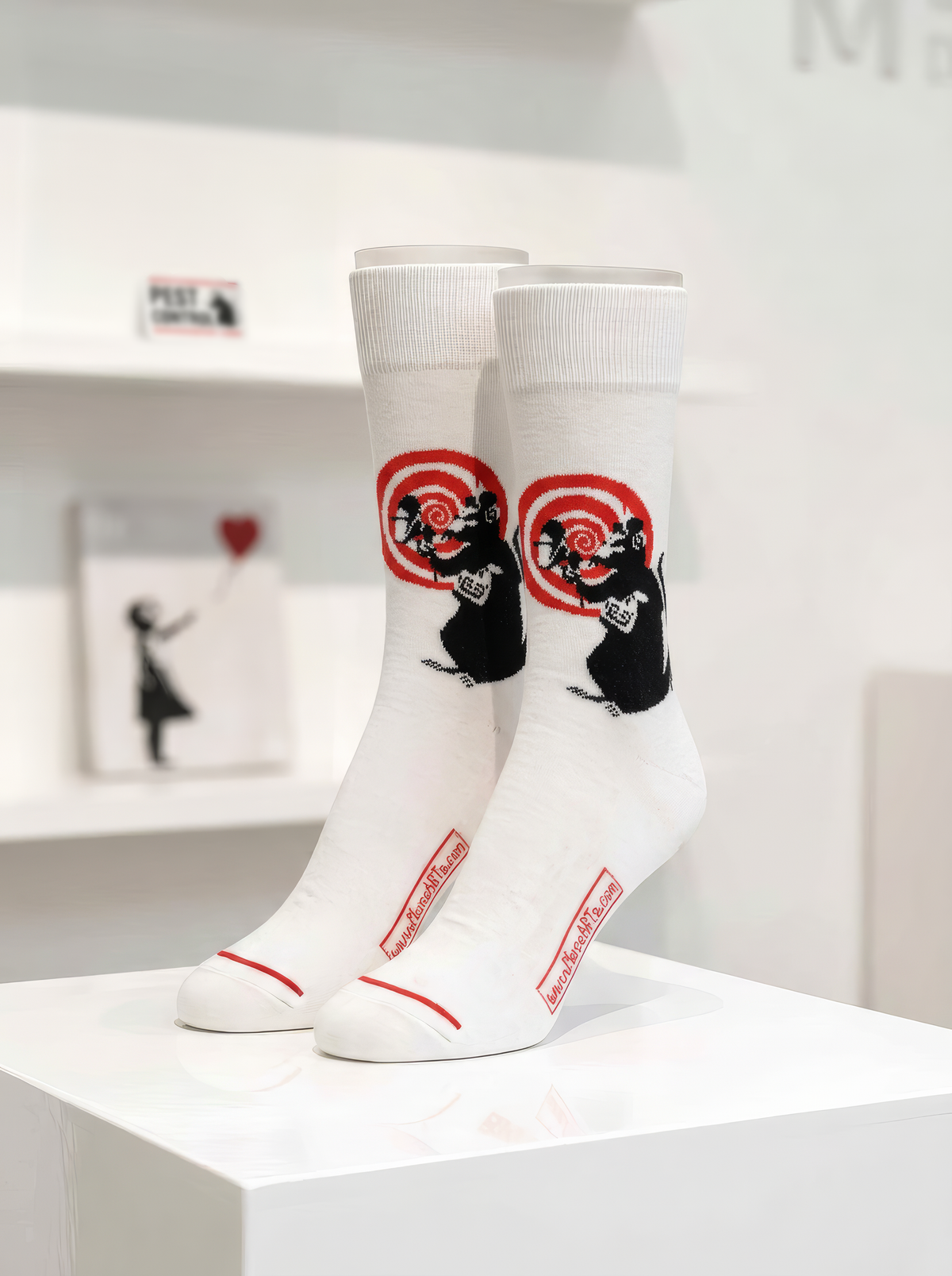 Pair of white Banksy-inspired Radar Rat art socks with black rat motifs and red circular details, displayed on a white plinth in a gallery-style setting.



