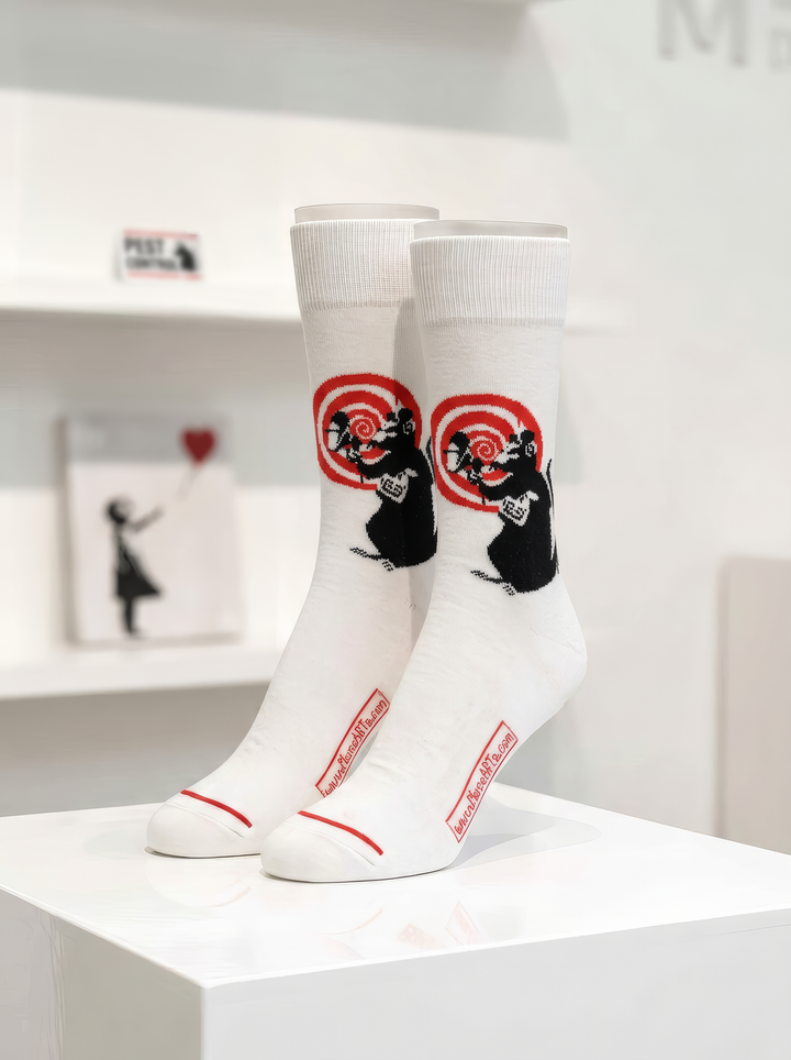 Pair of white Banksy-inspired Radar Rat art socks with black rat motifs and red circular details, displayed on a white plinth in a gallery-style setting.


