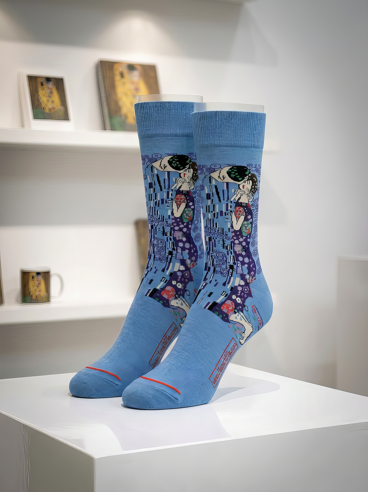 Pair of officially licensed blue Gustav Klimt The Kiss art socks displayed on a white gallery pedestal. Unisex art socks for men and women, suitable as a stylish gift for art lovers.