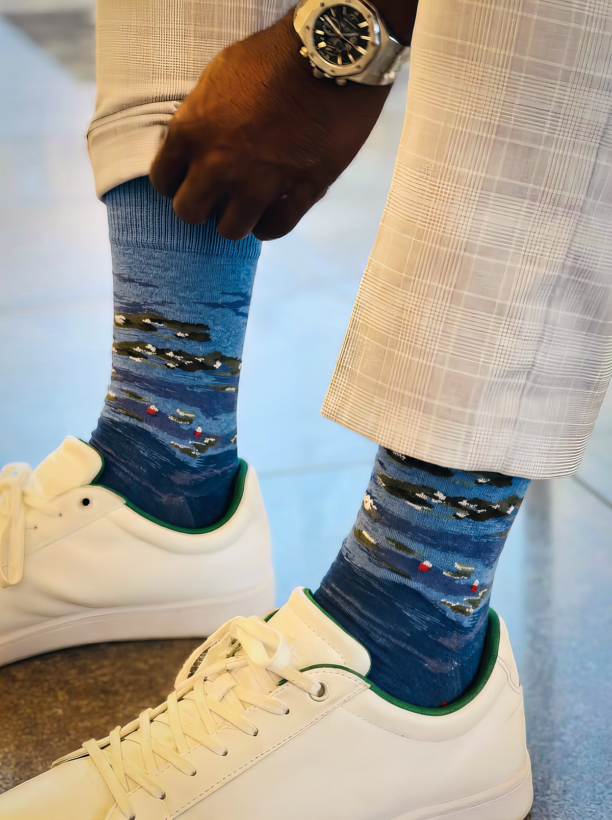 Close-up of a person adjusting blue XL socks featuring Claude Monet’s Water Lilies by Evening Light, styled with beige plaid trousers and white sneakers.