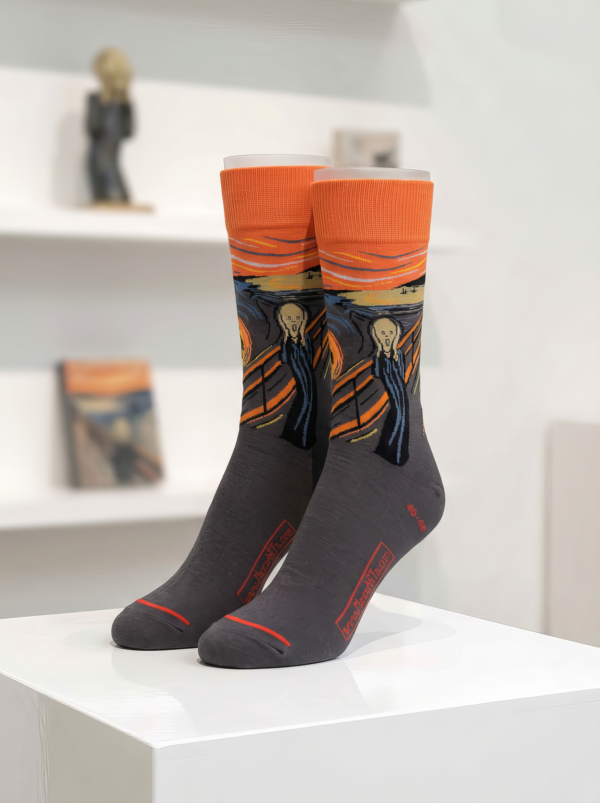 Dark Edvard Munch The Scream art socks with orange cuffs, displayed upright as a pair on a white plinth in a museum-style setting.
