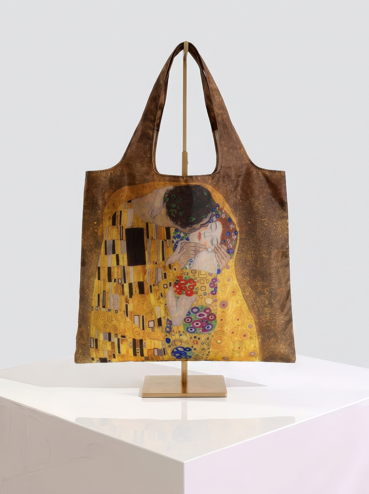 Foldable museum art shopper bag featuring The Kiss by Gustav Klimt, displayed on a gold stand against a white museum-style background from ArtyGifts
