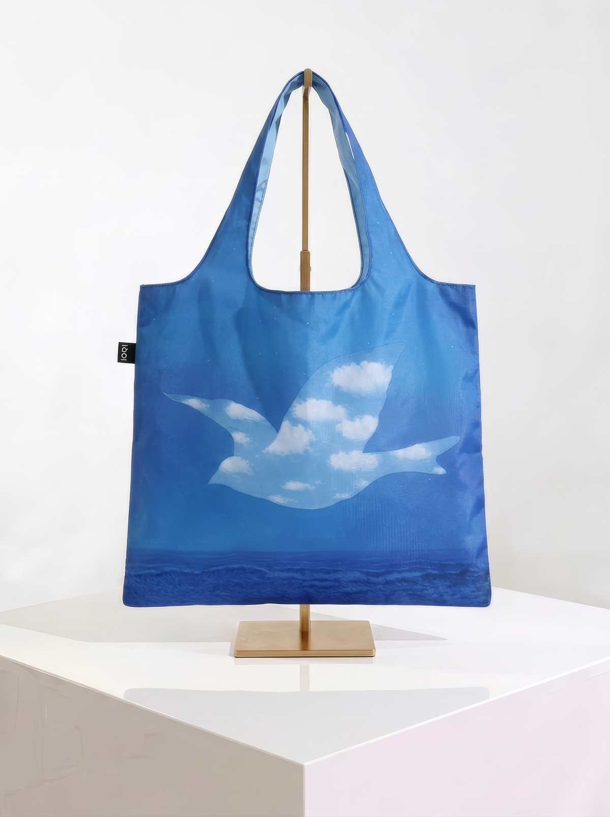 Foldable museum art shopper bag featuring The Promise by René Magritte, displayed on a gold stand against a white museum-style background from ArtyGifts
