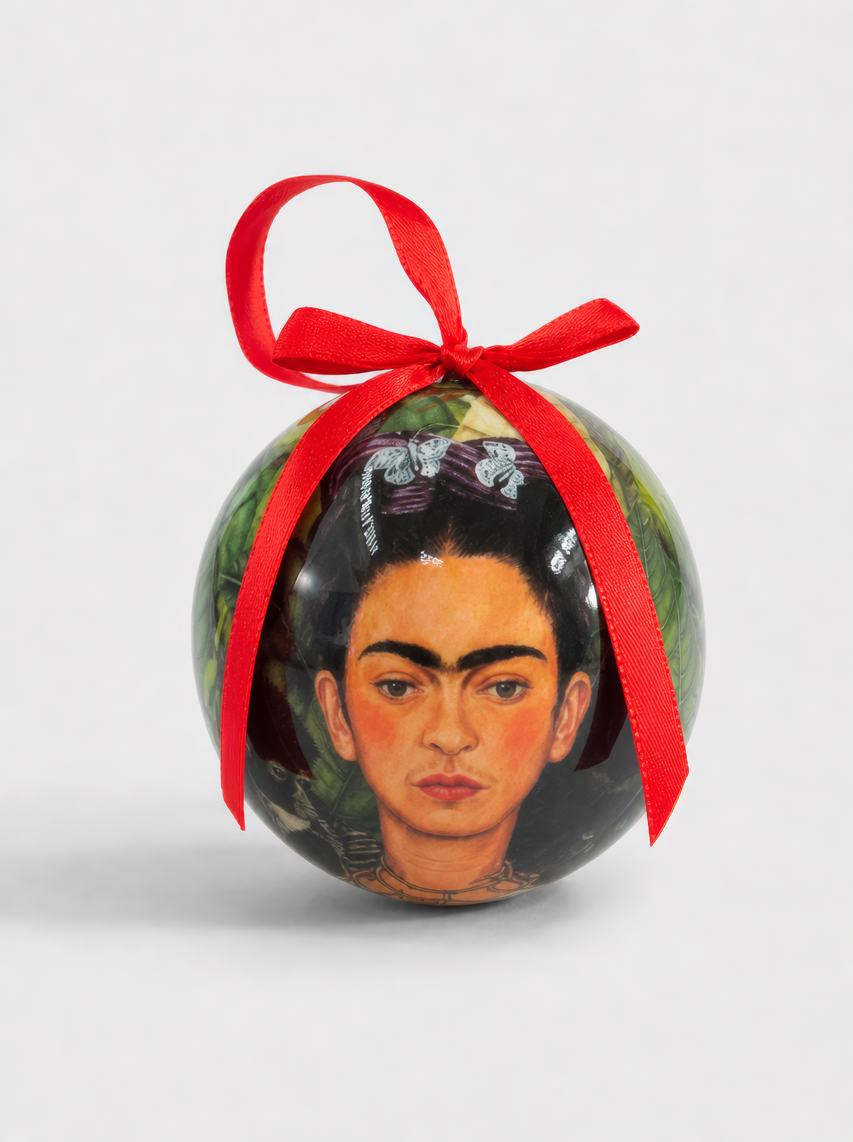 Close-up of a Frida Kahlo Self-Portrait Christmas bauble with red ribbon on a light grey background, showing Frida's iconic face with flower hair accessories and green jungle background – officially licensed museum art ornament, 7 cm diameter, unbreakable, in gift box. A unique Mexican art Christmas gift from ArtyGifts.
