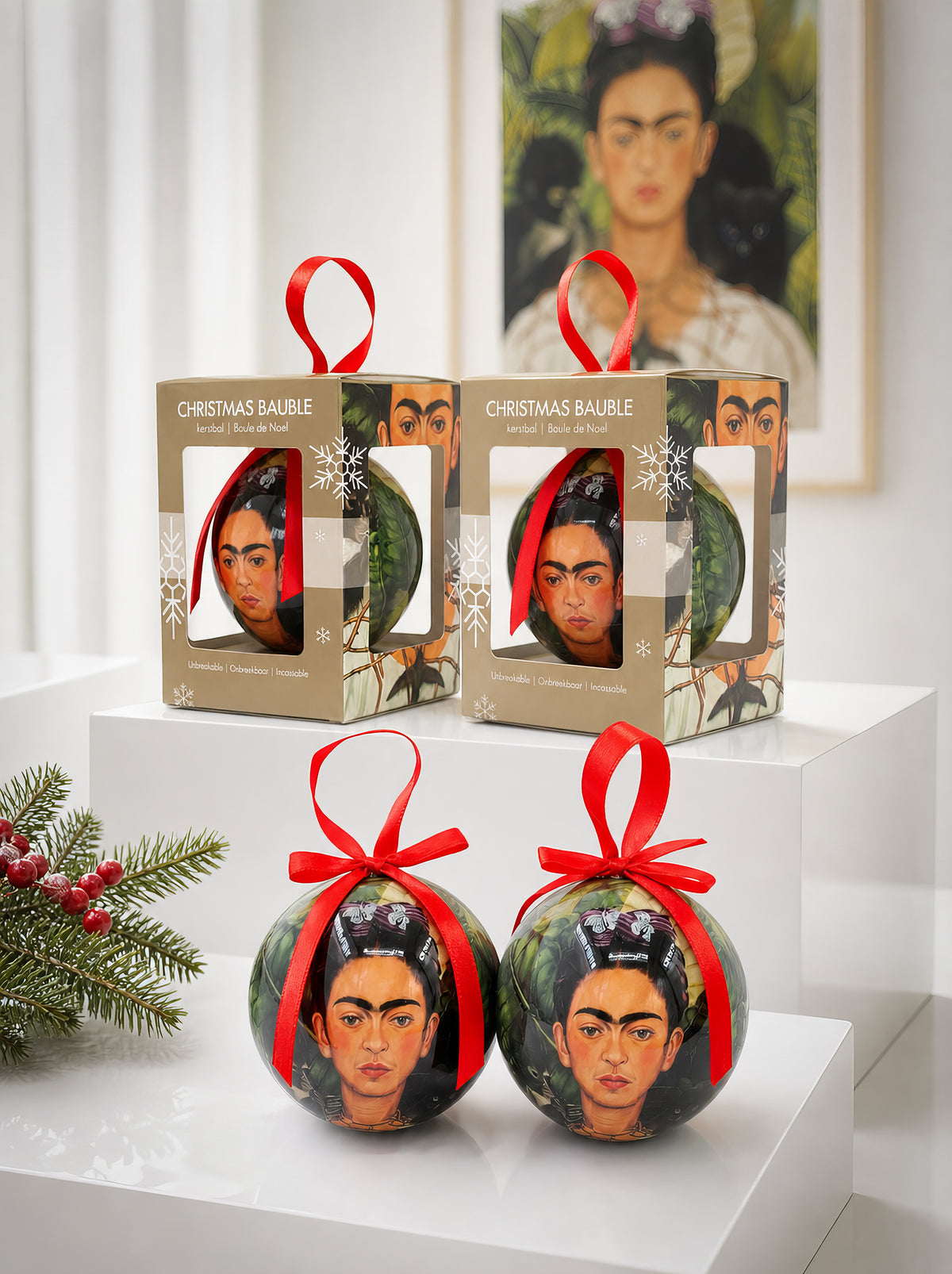 Two Frida Kahlo Self-Portrait Christmas baubles with red ribbons on a white pedestal next to their gift boxes, with a framed Frida Kahlo Self-Portrait print and pine branch with red berries in the background – officially licensed museum art ornament, 7 cm diameter, unbreakable. A unique Mexican art Christmas gift from ArtyGifts.