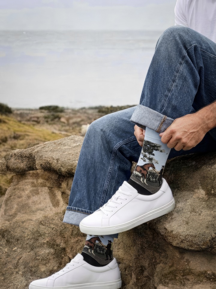 Person seated on coastal rocks wearing charcoal grey ArtyGifts horse socks with pale blue cuffs and white sneakers.

