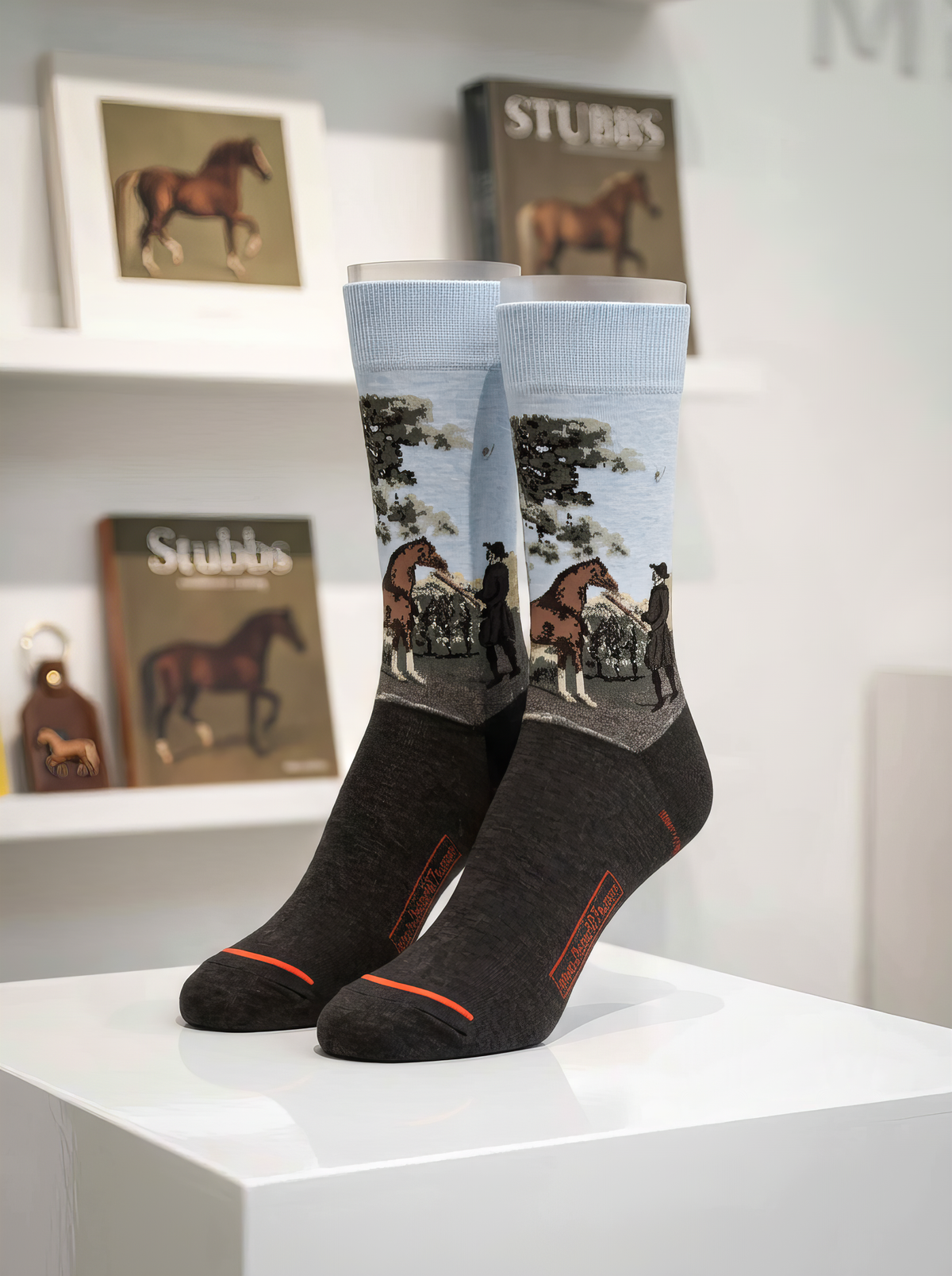 Pair of charcoal grey ArtyGifts socks with pale blue cuffs, featuring a George Stubbs horse scene, displayed on a white stand with Stubbs books and prints in the background.⠋