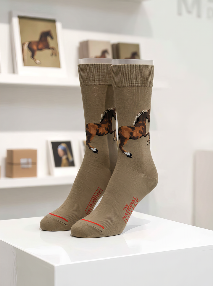 Pair of taupe art socks from ArtyGifts featuring George Stubbs’s horse artwork, displayed on a white pedestal in a museum shop.