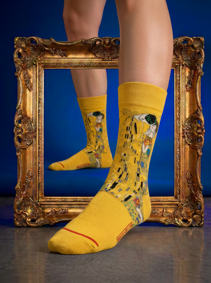 Person wearing officially licensed golden-yellow Gustav Klimt The Kiss art socks in front of an ornate gold mirror, showing the detailed woven artwork and red toe accent with a clear reflection. Unisex art socks for men and women, suitable as a stylish gift for art lovers.⠋