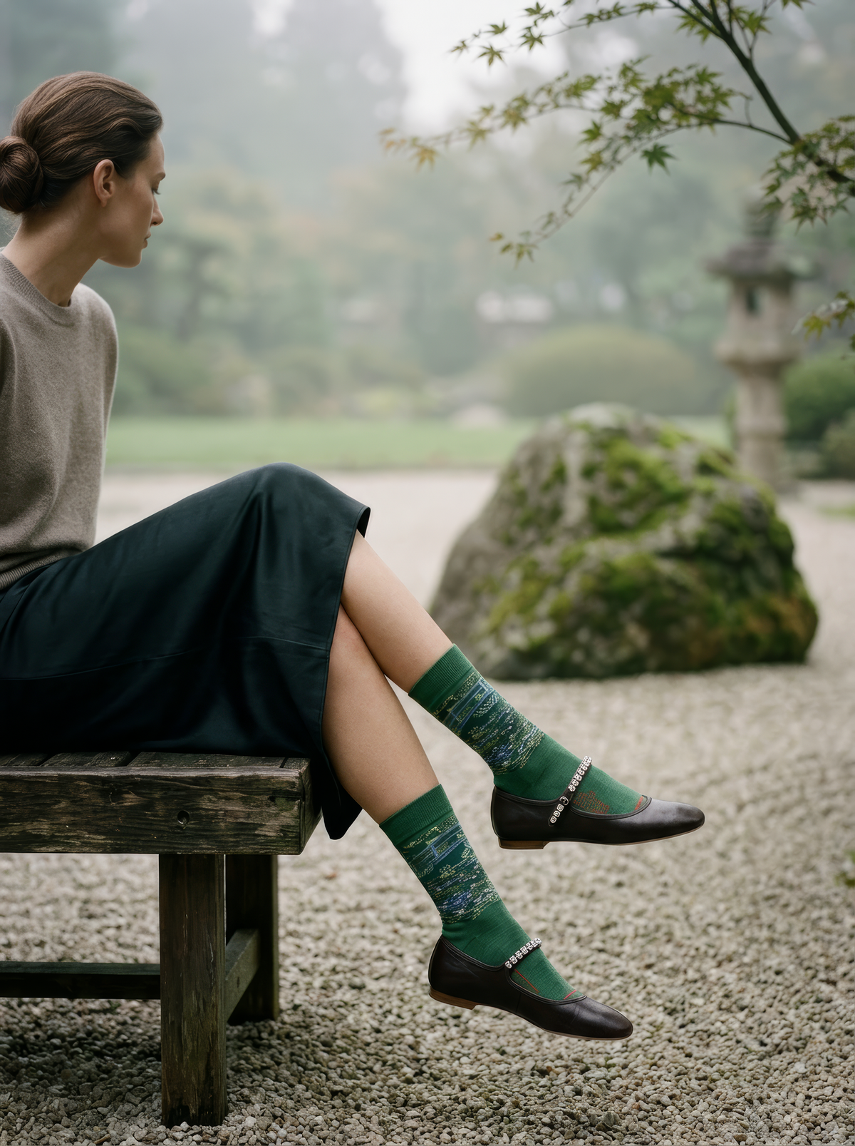 Person wearing green Claude Monet The Japanese Bridge art socks with dark clothing and dark shoes beside a garden landscape. For women and men.