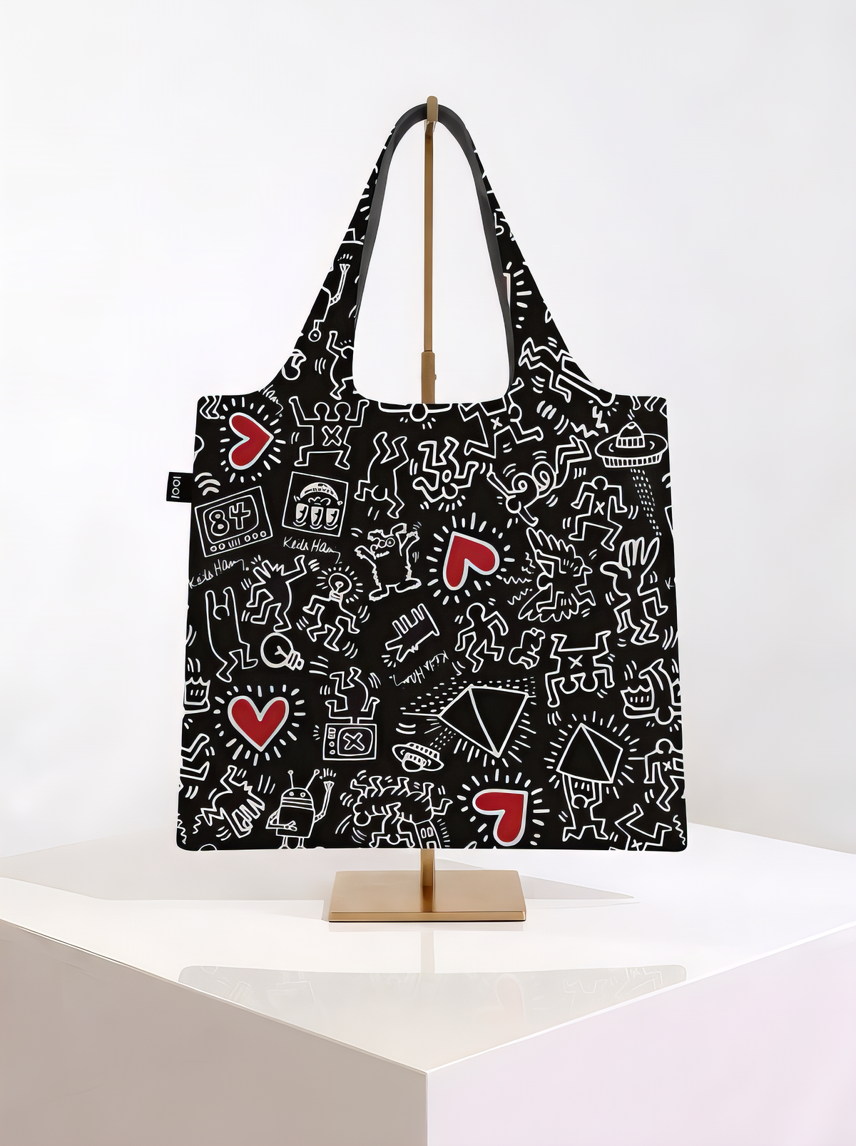 Black LOQI Keith Haring foldable shopper bag with white outlined figures and red hearts, hanging on a gold-coloured stand on a white pedestal.
