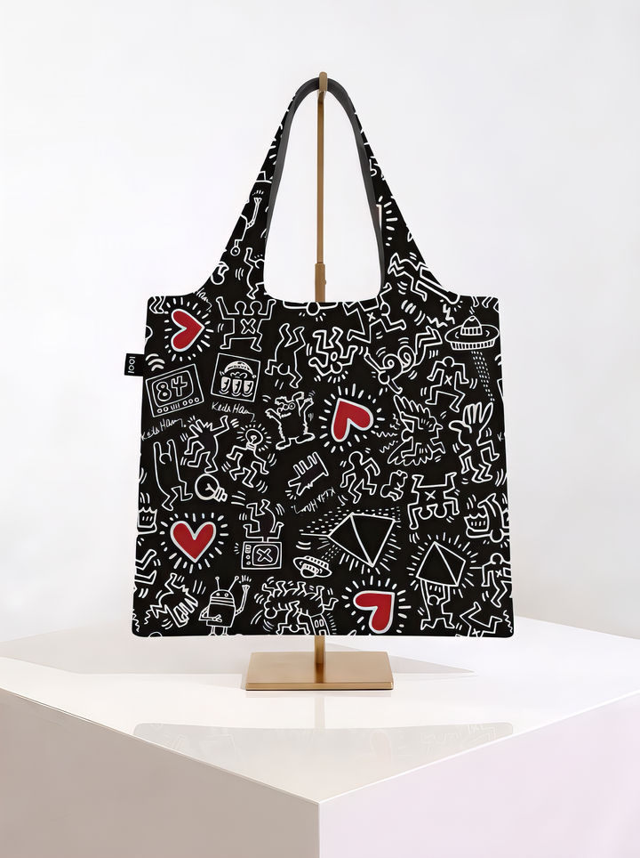 Black LOQI Keith Haring foldable shopper bag with white outlined figures and red hearts, hanging on a gold-coloured stand on a white pedestal.

