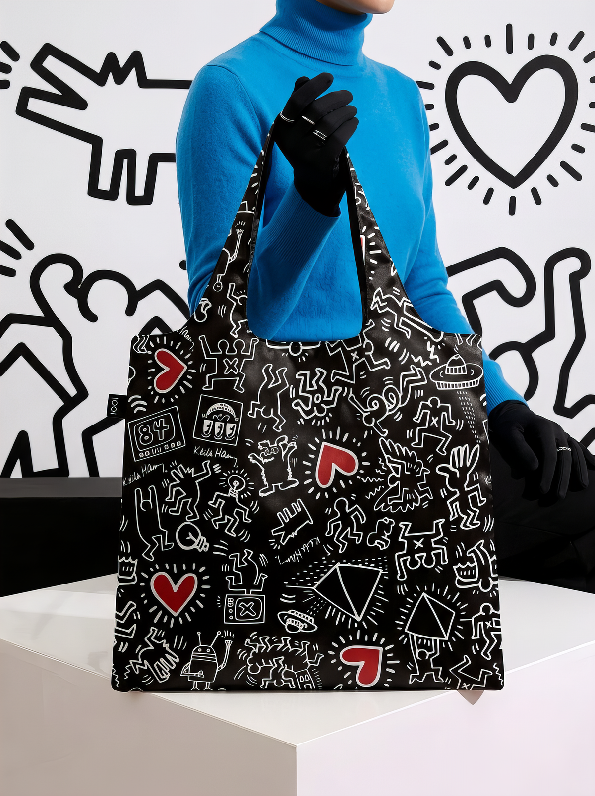 Person in a bright blue turtleneck and black gloves holding a LOQI Keith Haring shopper bag on a white pedestal, against a black-and-white illustrated wall.
