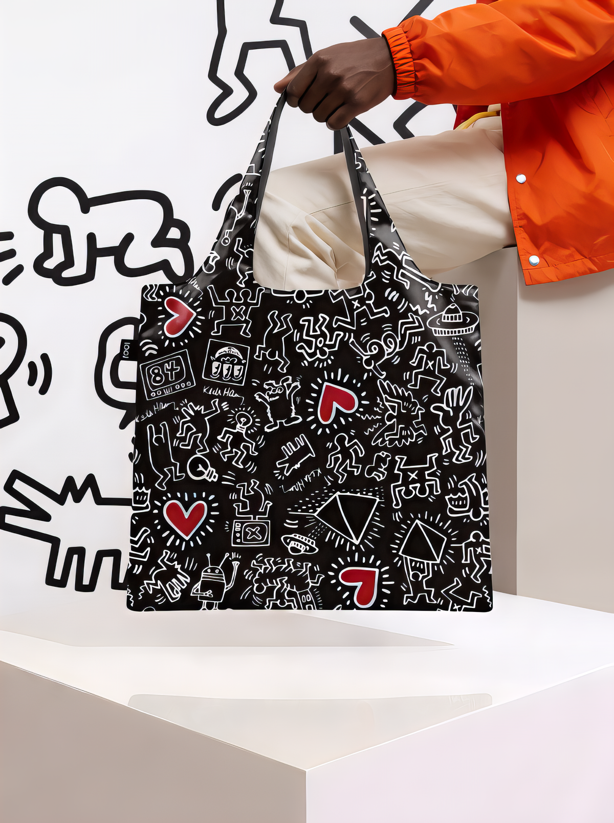Person in an orange long-sleeved top holding a LOQI Keith Haring Black Pattern shopper bag above a white pedestal, with outlined figures on the wall behind.
