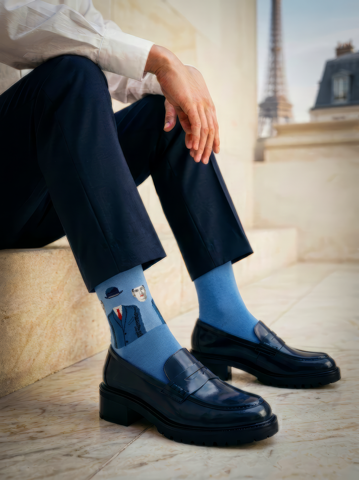 A seated person wears blue patterned art socks with cropped black trousers and chunky black shoes in a light interior.
