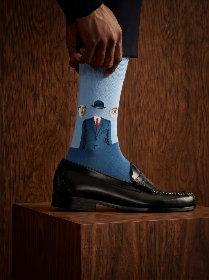 Close-up of a blue art sock featuring bowler-hatted figures, worn with a black leather shoe resting on a wooden plinth in a dimly lit setting.
