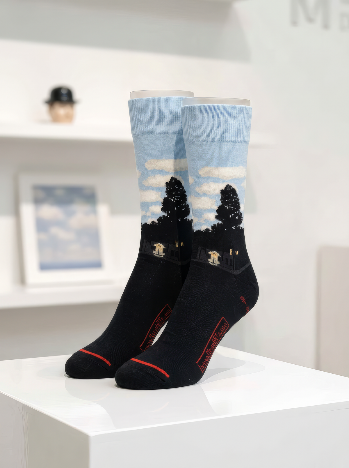 Pair of art socks with pale blue sky, white clouds and dark silhouettes above black feet, displayed upright on a white museum-style plinth.
