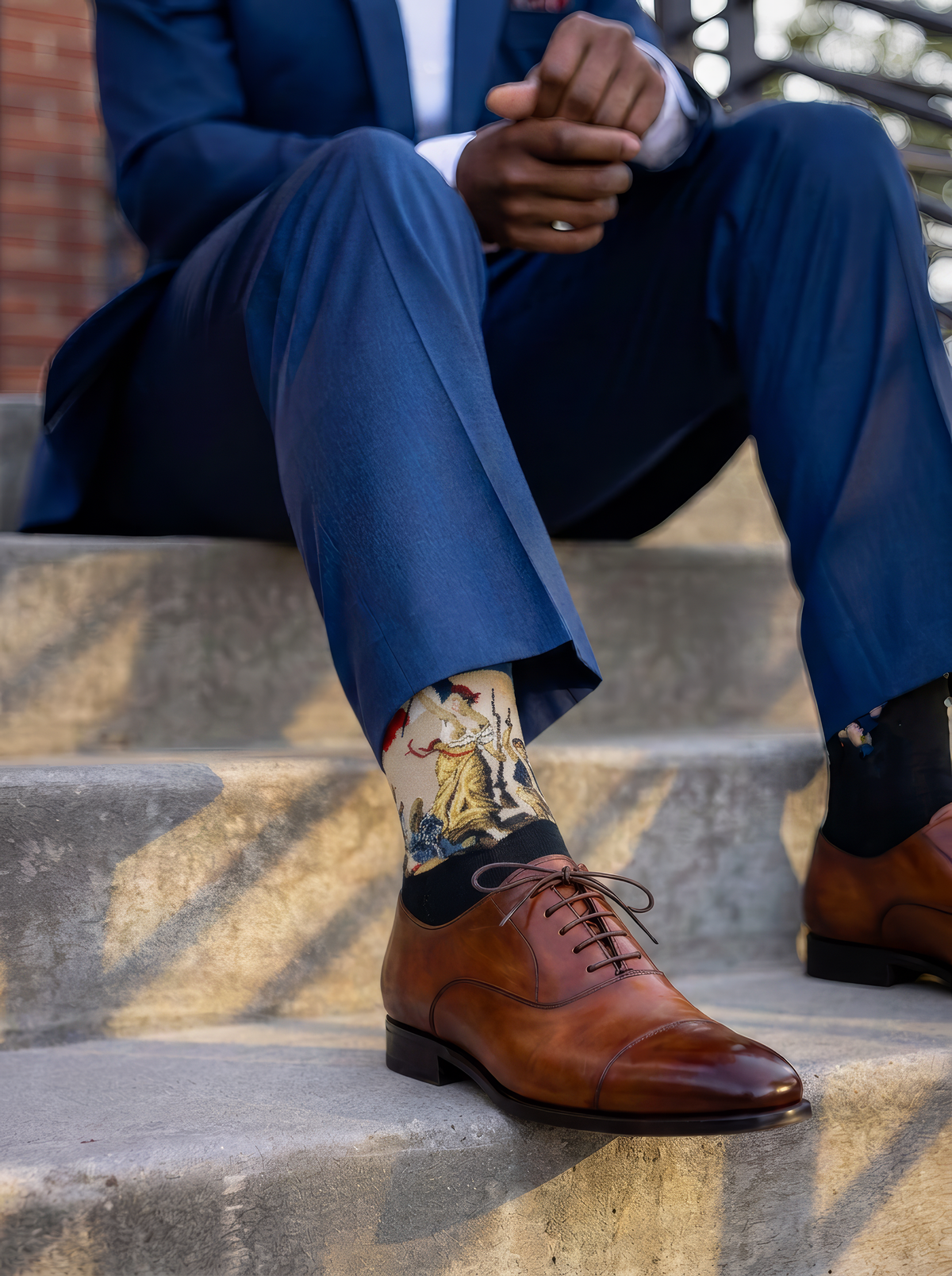 A seated person wears cream-patterned art socks with blue trousers and brown leather lace-up shoes in a light interior.
