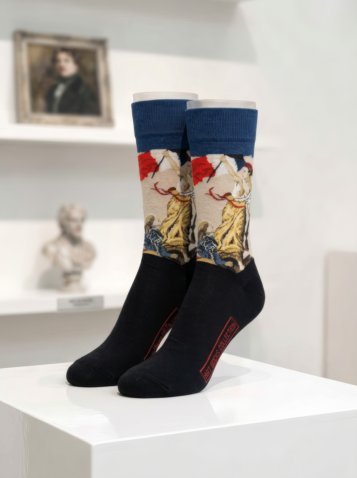 Pair of museum art socks with navy cuffs, a cream artwork panel with red and yellow details, and black feet, displayed on a white plinth.
