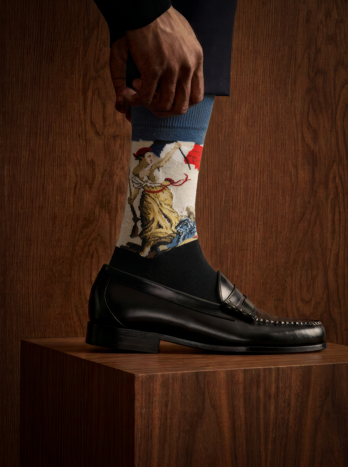 Close-up of a patterned art sock worn with a black leather shoe resting on a wooden plinth in a dimly lit setting.

