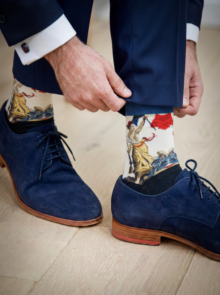 Close-up of a person adjusting an art sock above a navy lace-up shoe, showing the cream patterned section beneath dark trousers.
