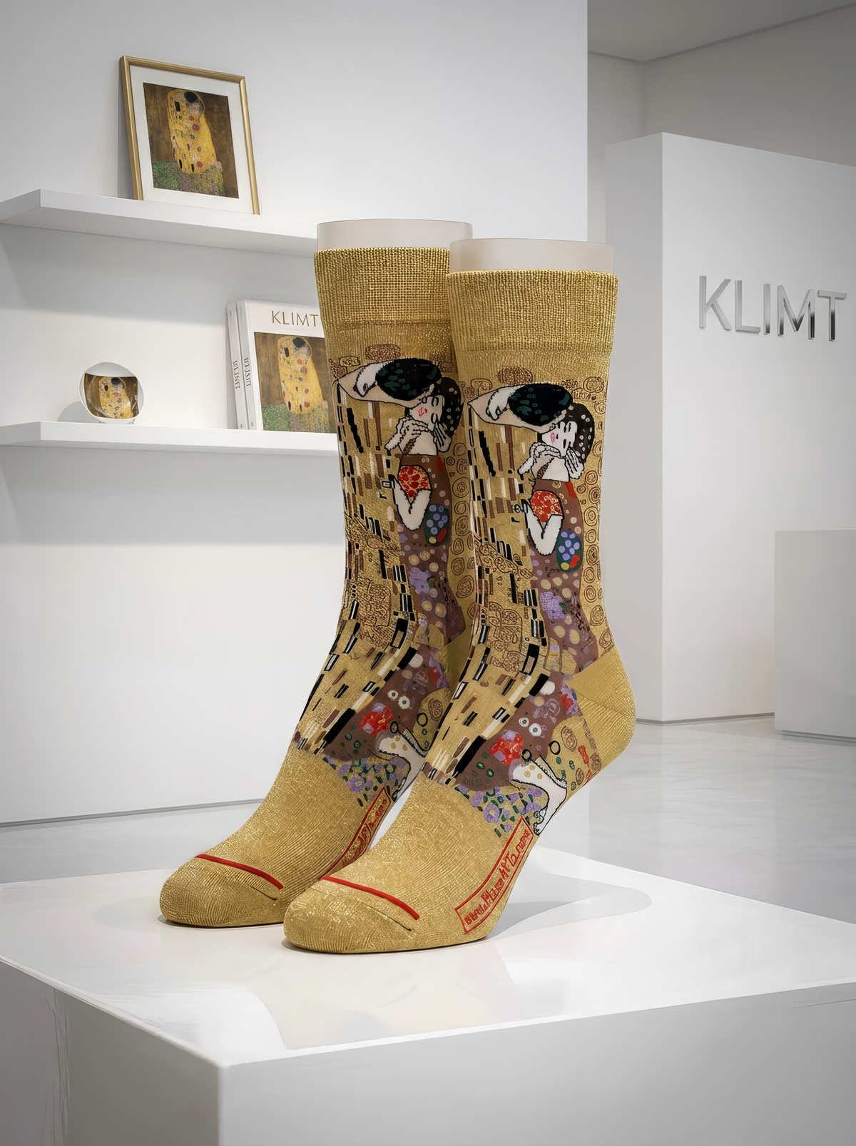 Pair of officially licensed gold Gustav Klimt The Kiss art socks displayed on a white gallery pedestal, showing the detailed woven artwork. Unisex art socks for men and women, suitable as a gift for art lovers.