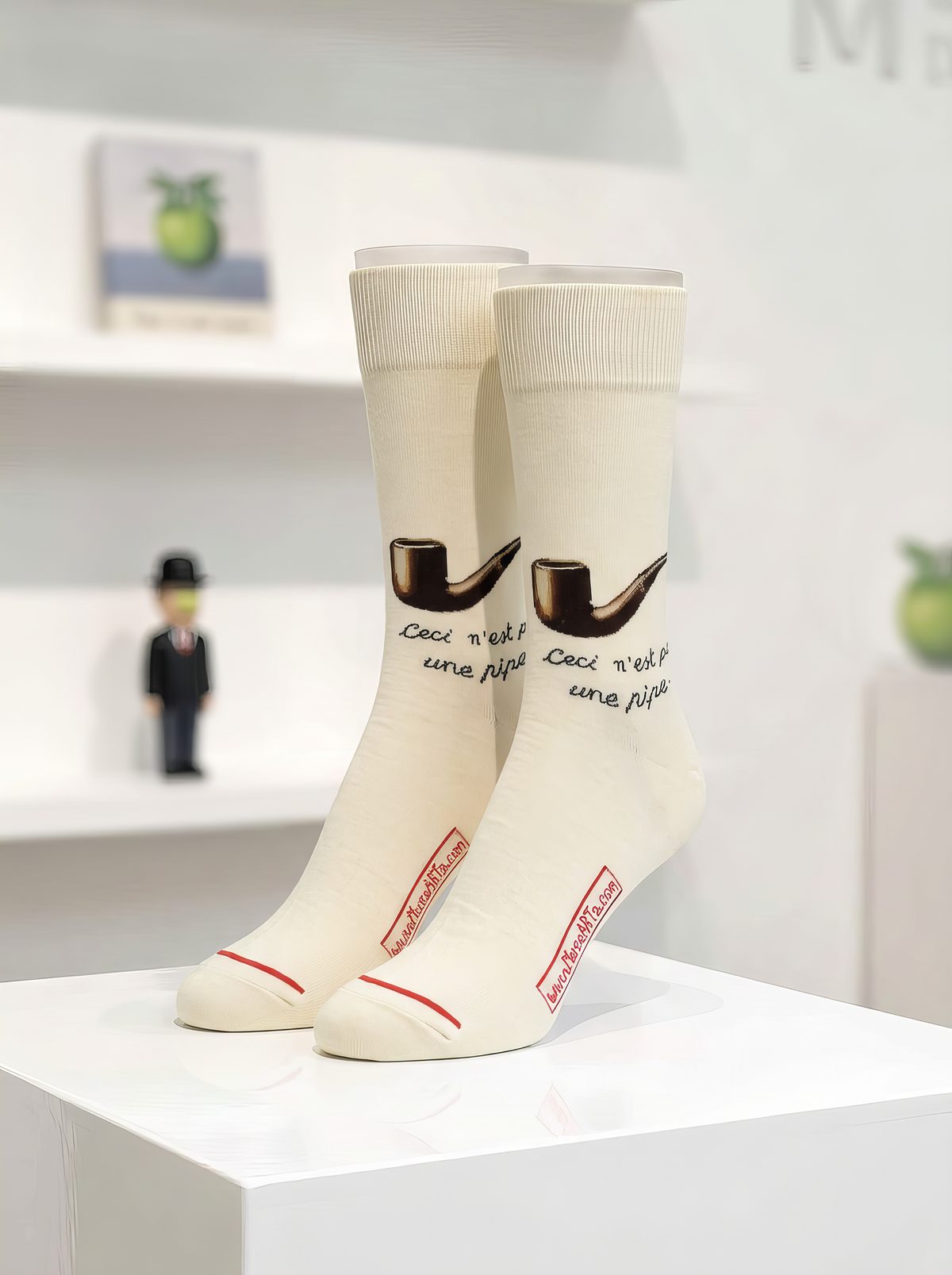 Cream René Magritte art socks with brown pipe motifs and “Ceci n’est pas une pipe” lettering, displayed on a white plinth in a museum-style setting.
