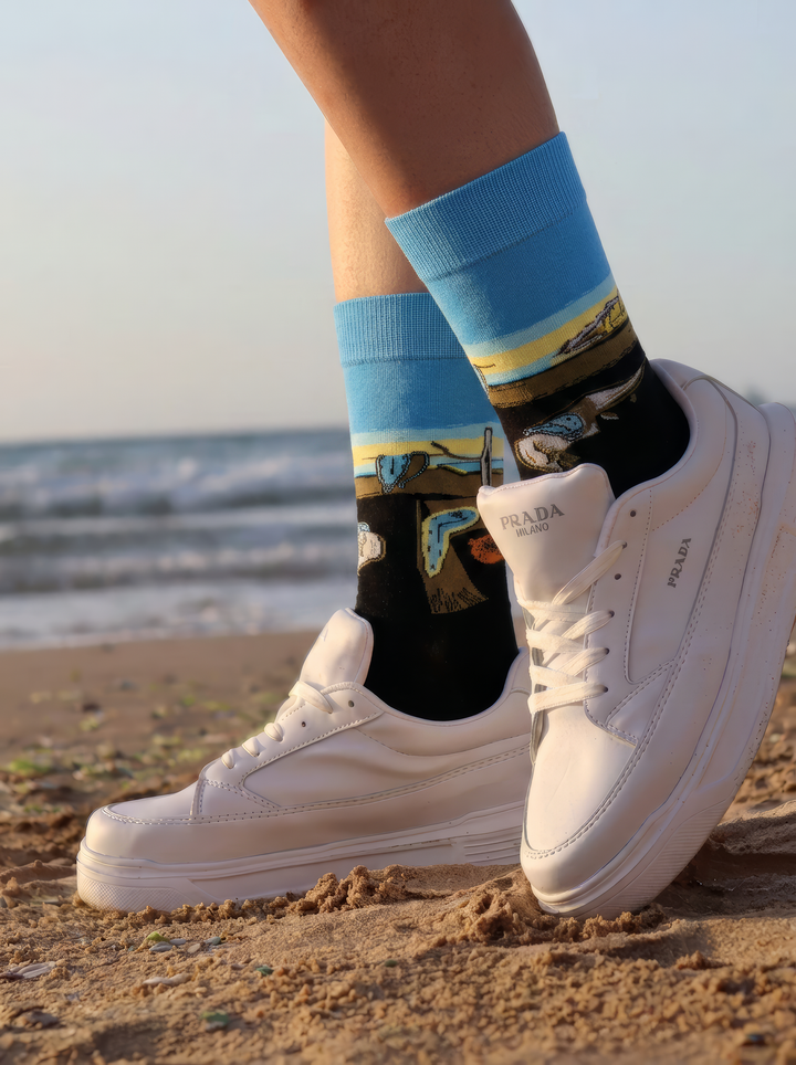 Pair of black XL socks with blue cuffs and yellow bands featuring Salvador Dalí artwork, worn with white sneakers on a sandy beach.
