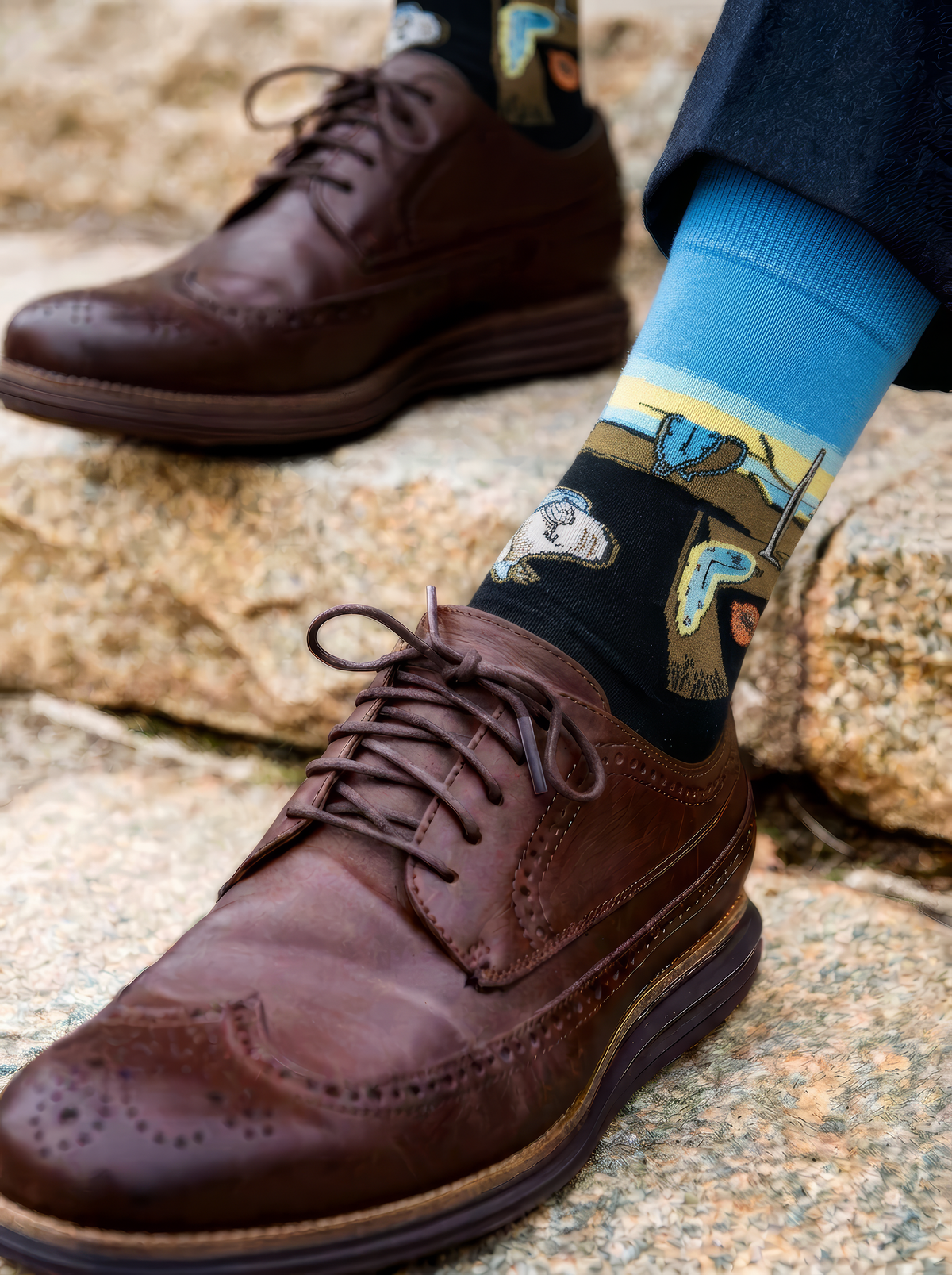 Close-up of a black XL crew sock with a blue cuff featuring Salvador Dalí artwork, paired with a brown brogue shoe; the matching sock and shoe appear in the background.