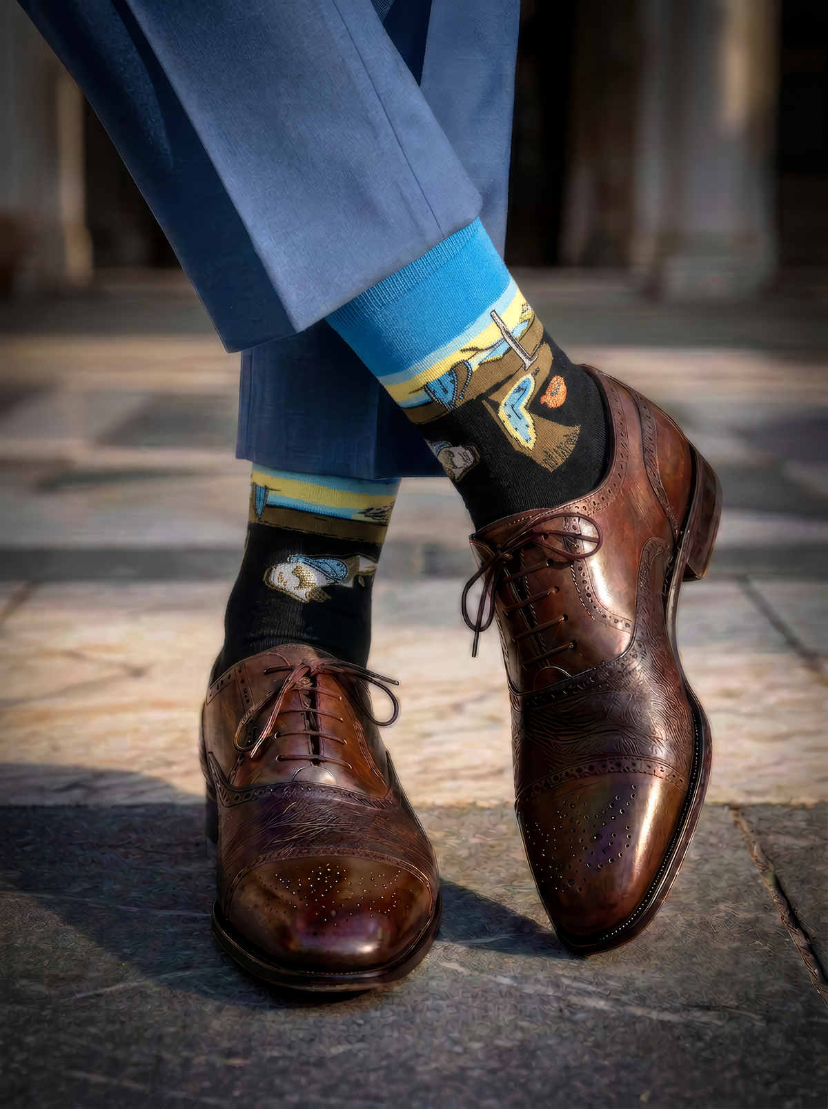 Close-up of a person in blue trousers wearing black XL socks with colourful Salvador Dalí artwork, styled with polished brown brogue shoes.⠋

